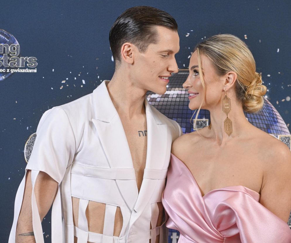 Hanna Żudziewicz i Jacek Jeschke, znani tancerze z programu „Taniec z gwiazdami”, uśmiechają się do siebie na tle logotypu show. Hanna w różowej sukni, Jacek w białej marynarce. Więcej o ich dramatycznych wspomnieniach z pożaru auta przed ślubem przeczytasz na naszym portalu.