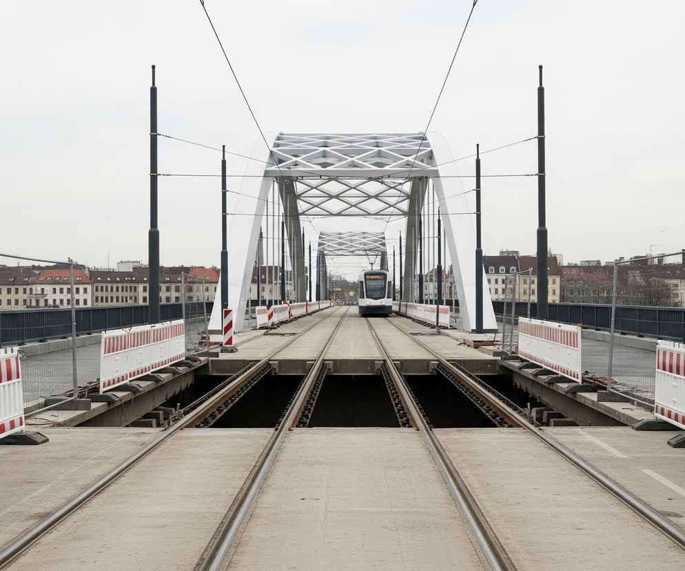 Na pierwszym planie widoczne są dwa tory tramwajowe, biegnące przez most o stalowej konstrukcji kratownicowej, pomalowanej na biało, z torami ułożonymi na betonowej nawierzchni. Po obu stronach torów stoją biało-czerwone barierki budowlane, a dalej, wzdłuż krawędzi mostu, rozciąga się metalowe ogrodzenie. W oddali, na końcu mostu, widać nadjeżdżający biało-niebieski tramwaj. Za mostem rozciąga się panorama miasta z budynkami o ceglanych dachach, a całe niebo jest jasnoszare.