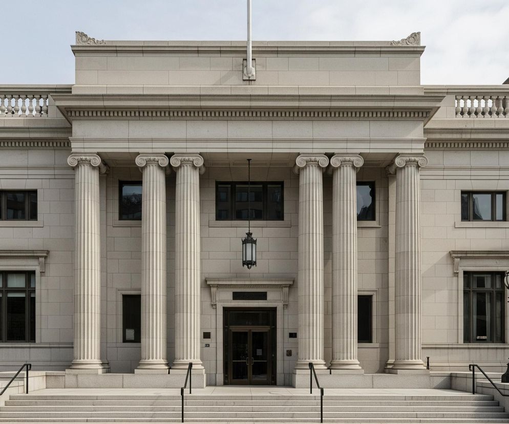 Centralną część obrazu zajmuje monumentalna, jasnoszara fasada budynku z elementami architektury klasycznej. Przed wejściem głównym znajduje się sześć wysokich, żłobionych kolumn z jońskimi kapitelami, wspierających gzyms i balustradę z tralek. Wejście stanowią dwuskrzydłowe, ciemne drzwi, nad którymi wisi ozdobna, czarna latarnia. Schody z szerokimi, szarymi stopniami prowadzą do wejścia, otoczone niskimi murkami z czarnymi poręczami, a po bokach schodów stoją dwie identyczne, czarne latarnie uliczne. W tle po prawej stronie widoczny jest fragment ceglanego budynku z wieloma oknami, a nad całością rozciąga się jasne, zachmurzone niebo.