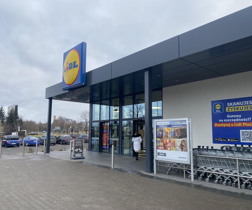 Zewnętrzny widok na wejście do sklepu Lidl, z charakterystycznym niebiesko-żółtym logo. Obok wejścia rząd wózków sklepowych oraz plakat informujący o promocjach na karpia. Tło stanowi pochmurne niebo i parking. Czytaj więcej o świątecznych promocjach na Super Biznes.