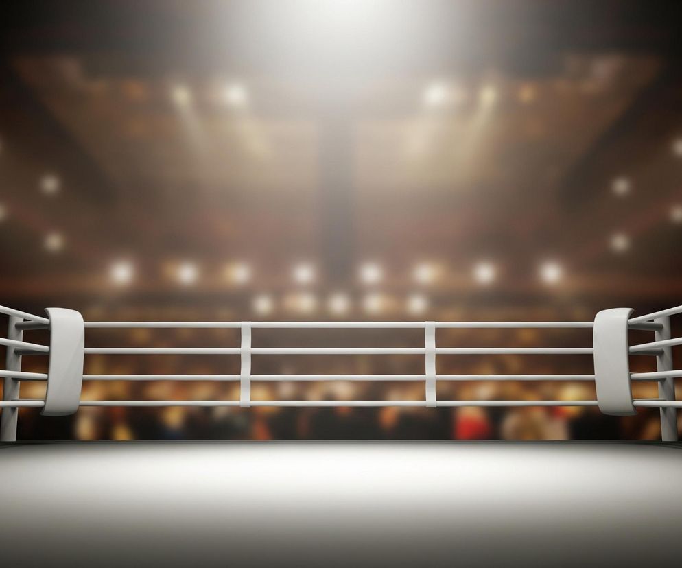 Na pierwszym planie widoczny jest szary ring bokserski z trzema białymi linami otaczającymi go. Liny są przymocowane do czterech pionowych słupków, z których dwa boczne są częściowo widoczne. Tło jest rozmyte, przedstawiające ciemną przestrzeń z licznymi, jasnymi, rozmazanymi światłami w odcieniach żółci i bieli, symulującymi publiczność i oświetlenie areny. Górna część tła jest najjaśniejsza, z intensywnym, rozświetlonym obszarem na środku.