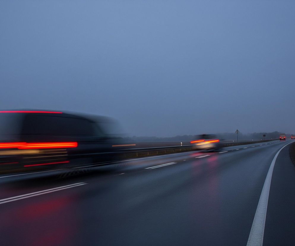Pasmowa droga asfaltowa biegnie przez centralną część kadru, z lewej strony widoczne są dwa białe pasy oddzielające pasy ruchu, a po prawej barierka ochronna. Dwa rozmazane pojazdy jadą w lewo, jeden bliżej widza, drugi dalej, ich tylne czerwone światła tworzą smugi światła. Niebo jest jednolicie szare, a horyzont pokrywa mgła, która rozmywa widok pól i drzew. W oddali, po prawej stronie drogi, widać znak drogowy na tle ciemnych drzew.