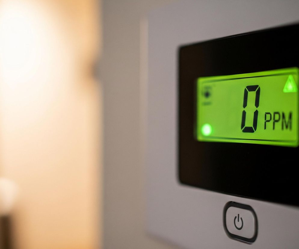 Zbliżenie ukazuje elektroniczny wyświetlacz z jasnym, zielonym tłem, na którym widnieje duża cyfra 0 oraz tekst PPM. W lewym górnym rogu wyświetlacza znajduje się mała, zielona ikona przypominająca głośnik, a w prawym górnym rogu zielony, świecący trójkąt. Poniżej wyświetlacza, na białym panelu urządzenia, widoczny jest przycisk włączania/wyłączania z symbolem zasilania. Lewa strona tła jest rozmyta, ukazując ciepłe, jasne odcienie brązu i beżu, zaś prawa strona tła jest mniej oświetlona, utrzymana w szarych tonacjach.