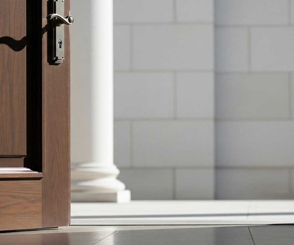 Prawie całkowicie otwarta, ciemnobrązowa drewniana framuga drzwi widoczna jest po lewej stronie kadru, z mosiężną klamką o prostym, podłużnym kształcie w górnej części. Na powierzchni drzwi pada cień klamki, tworząc falisty wzór. Za drzwiami, w głębi, znajduje się słabo oświetlony obszar z białymi, kwadratowymi płytkami na ścianie i fragmentem białej kolumny pośrodku. Dół kadru wypełnia ciemna, błyszcząca podłoga, odbijająca światło zza drzwi.