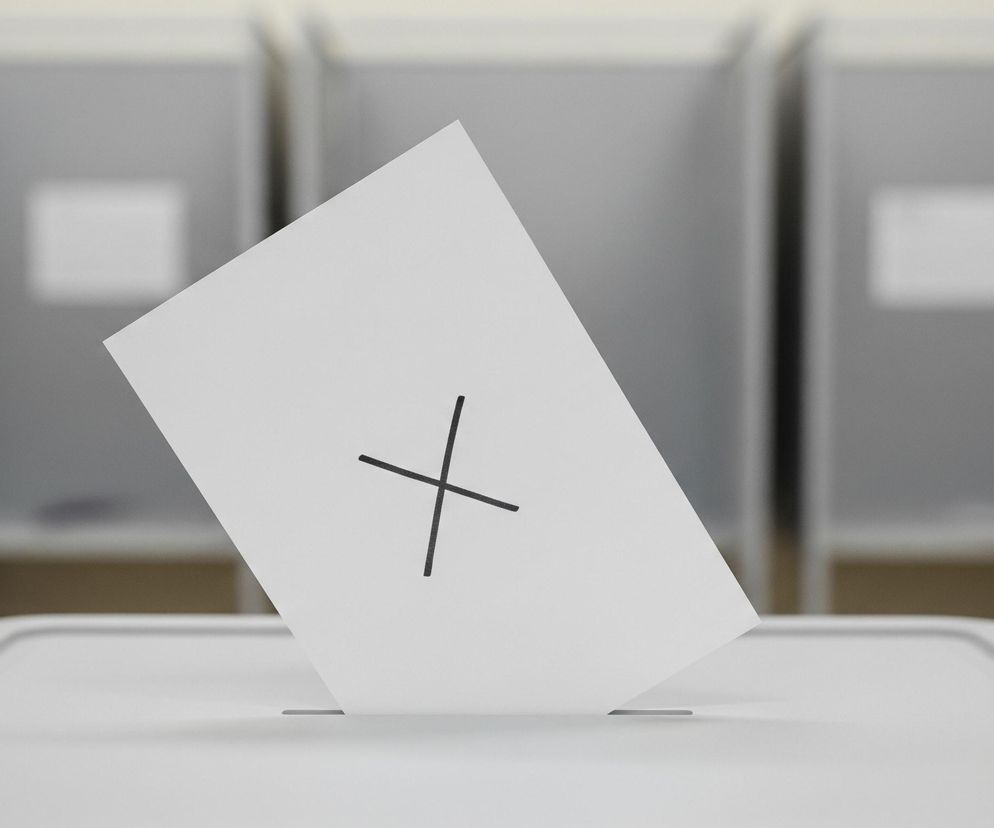 Biała karta do głosowania z dużym, czarnym znakiem X jest wsuwana do szczeliny w białej urnie wyborczej. Karta jest lekko pochylona w prawo, a jej dolna część znika w otworze urny. Tło stanowią trzy szare kabiny do głosowania, niewyraźne, z widocznymi wewnątrz białymi prostokątami symbolizującymi okna lub otwory. Cała scena utrzymana jest w jasnych, neutralnych barwach.