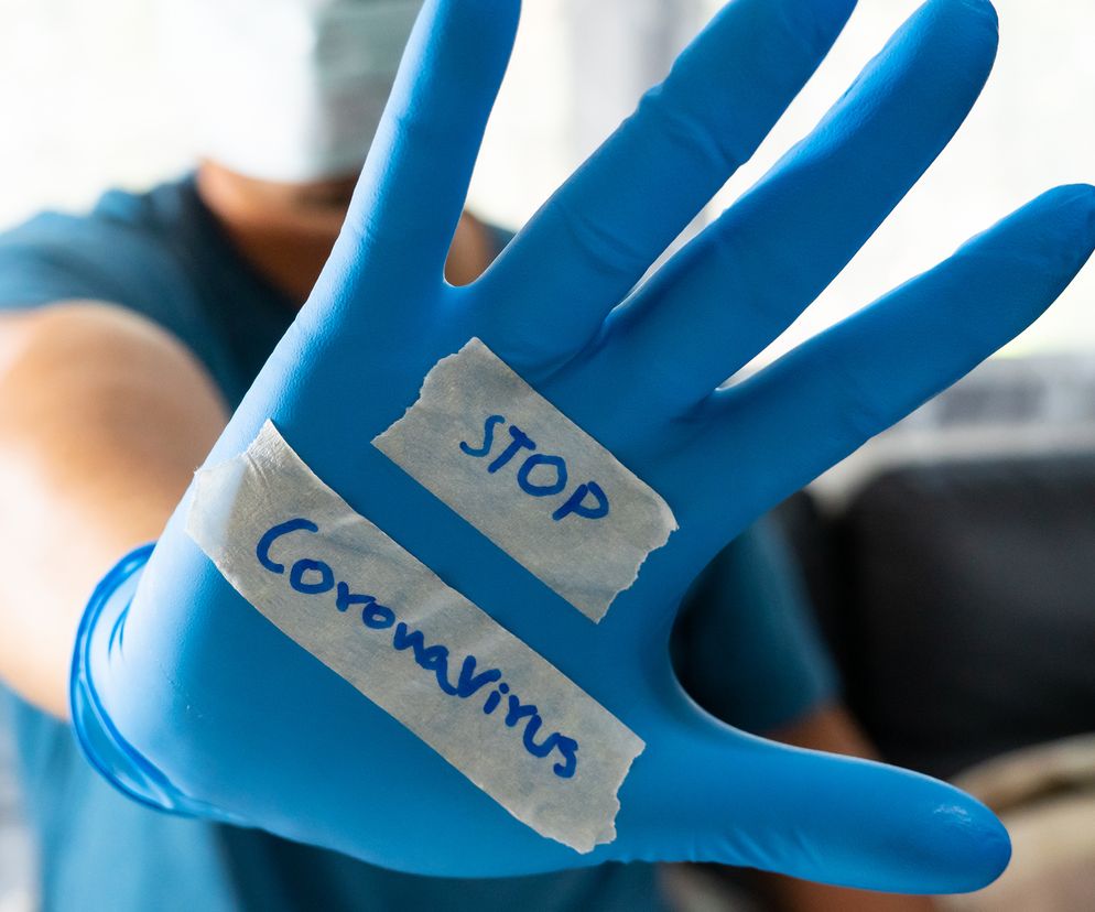Osoba w niebieskiej koszulce i masce ochronnej z ręką w niebieskiej rękawiczce wyciągniętą w kierunku odbiorcy. Na wewnętrznej stronie dłoni, na dwóch kawałkach taśmy, widnieją napisy STOP i Coronavirus, symbolizujące walkę z pandemią, o czym przeczytasz więcej na Poradnik Zdrowie.