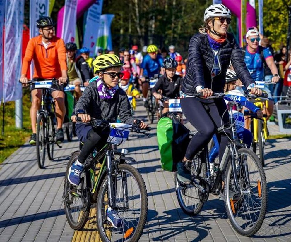 Rodzina i rowerzyści w kaskach, okularach przeciwsłonecznych i sportowych strojach na Rajdzie Rowerowym w Parku Śląskim. Uczestnicy jadą po wybrukowanej ścieżce, widoczne zielone trawniki i flagi. Więcej o wydarzeniu przeczytasz na Super Biznes.