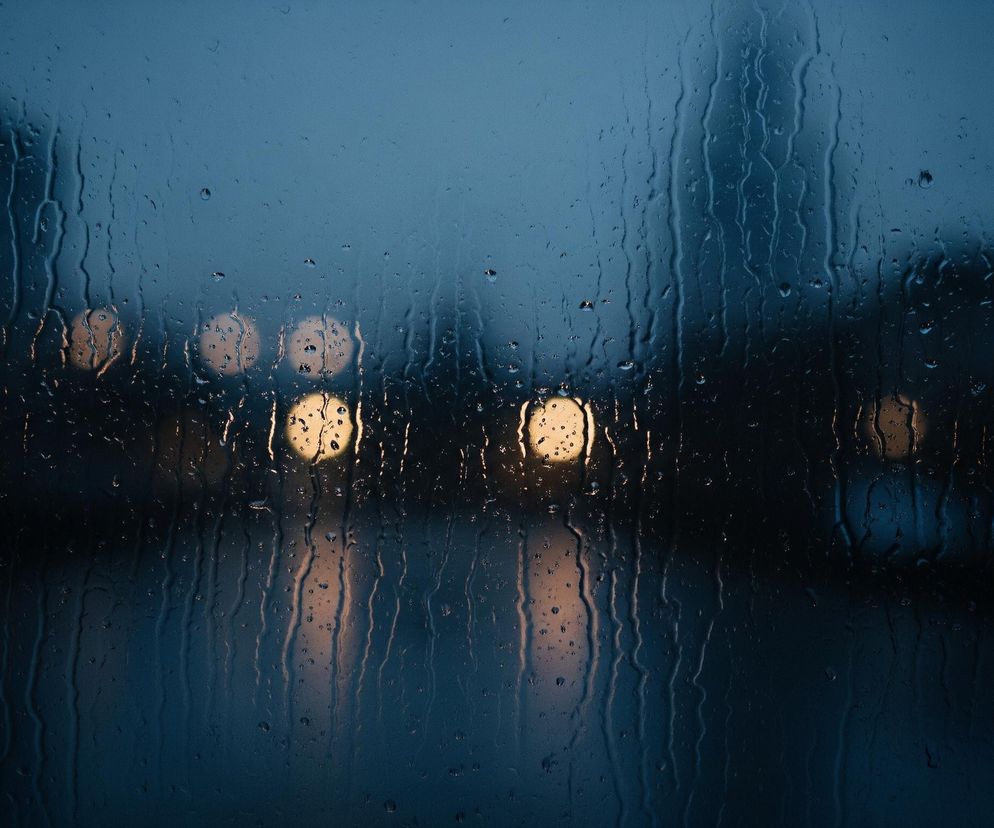 Szyba pokryta jest licznymi kroplami wody i smugami, co sugeruje deszcz lub inną formę wilgoci. W tle widać rozmazane, jasne okręgi w ciepłych odcieniach żółci i pomarańczu, prawdopodobnie światła uliczne lub inne źródła światła, które są zniekształcone przez wodę na szkle. Otoczenie tła jest ciemne, w dominujących odcieniach granatu i czerni, co wskazuje na wieczorową porę lub noc.