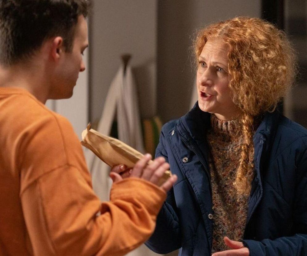 Kobieta o rudych, kręconych włosach, ubrana w granatową kurtkę i wzorzysty sweter, rozmawia z mężczyzną w pomarańczowej bluzie, który trzyma w dłoniach podłużny pakunek. Zdjęcie ilustruje scenę z serialu Barwy szczęścia, opisaną w artykule Super Seriale.