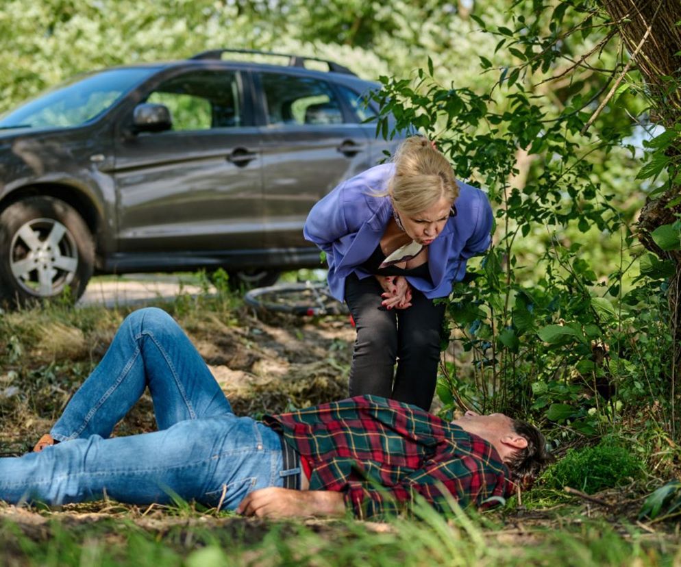 Kobieta pochylająca się nad leżącym mężczyzną, oboje na tle drzew i samochodu na poboczu drogi, scena z serialu Na Wspólnej, gdzie Boska (Sylwia Wysocka) ratuje Edwarda (Wojciech Skibiński). Więcej informacji o serialu Na Wspólnej znajdziesz na Super Seriale.
