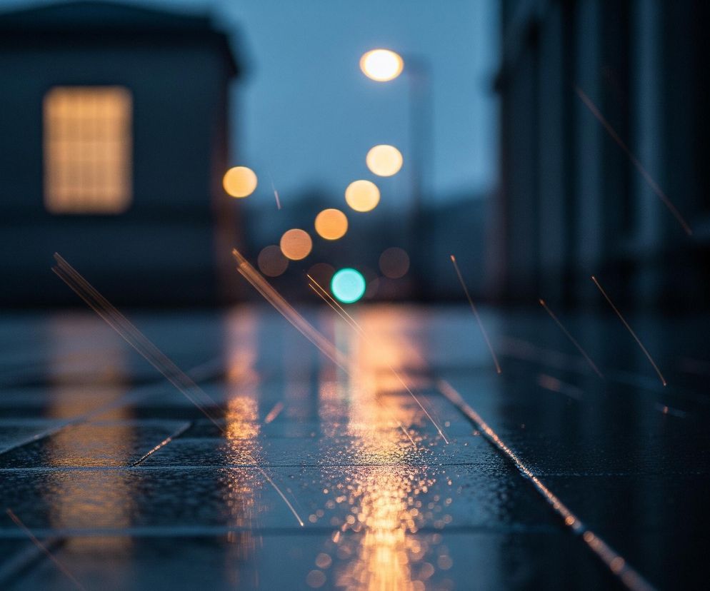 Zbliżenie na mokry, ciemny chodnik, odbijający światła latarni. Liczne krople wody na jego powierzchni mienią się żółto-pomarańczowymi i zielonkawymi refleksami, rozproszonymi liniami światła. W tle widoczne są rozmyte budynki o ciemnej fasadzie oraz szereg okrągłych, rozmytych świateł ulicznych, głównie pomarańczowych i jedno turkusowe. Niebo jest ciemnoniebieskie.