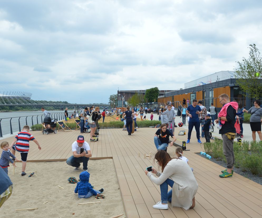 Ludzie, w tym dzieci, bawią się na Bulwarach Wiślanych w Warszawie, na tle rzeki i Mostu Świętokrzyskiego oraz Stadionu Narodowego. Mężczyzna w białej czapce i kobieta w beżowym swetrze robią zdjęcia telefonami, uwieczniając pożegnanie lata. Więcej o wydarzeniach kulturalnych przeczytasz na Poradnik Zdrowie.