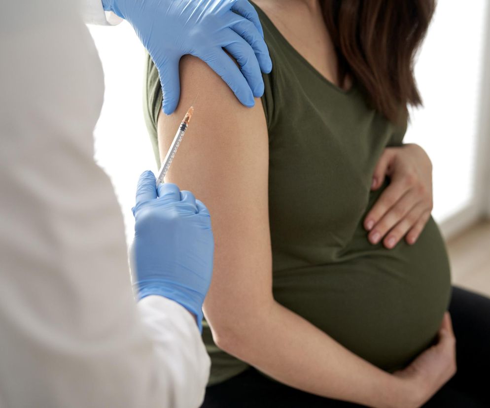Kobieta w ciąży w zielonej koszulce, z ręką na brzuchu, otrzymuje szczepienie w ramię od osoby w białym fartuchu i niebieskich rękawiczkach. Igła strzykawki jest wprowadzana w skórę. Więcej o szczepieniach w ciąży przeczytasz na Poradnik Zdrowie.