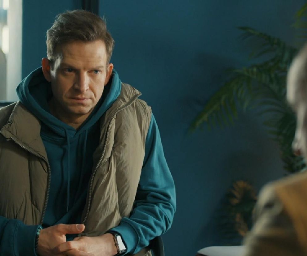 Michał Rolnicki jako Łukasz Sadowski w Barwach szczęścia, ubrany w turkusową bluzę z kapturem i beżową kamizelkę, siedzi podczas poważnej rozmowy z tyłu głowy widać postać Kornela (Jakub Bohosiewicz). Sprawdź streszczenia odcinków na Super Seriale.