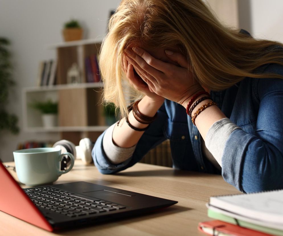 Zestresowana kobieta w niebieskiej koszuli jeansowej i jasnoszarej bluzce, zakrywająca twarz dłońmi, siedzi przy laptopie, obok filiżanka i słuchawki. Symbolizuje obawy przed utratą pracy przez AI. Dowiedz się więcej o tym na portalu Super Biznes.
