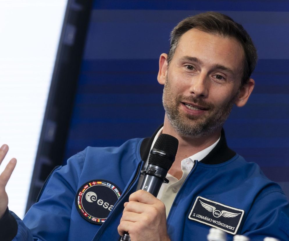 Sławosz Uznański-Wiśniewski, polski astronauta, uśmiecha się, trzymając mikrofon. Ma na sobie niebieską kurtkę z naszywkami ESA i flagami, w tym z imieniem i nazwiskiem. Gestykuluje lewą ręką podczas wystąpienia na 64. Forum Ekonomicznym w Karpaczu, gdzie dzieli się doświadczeniami z misji kosmicznej. Więcej na ten temat można przeczytać na portalu Super Biznes.