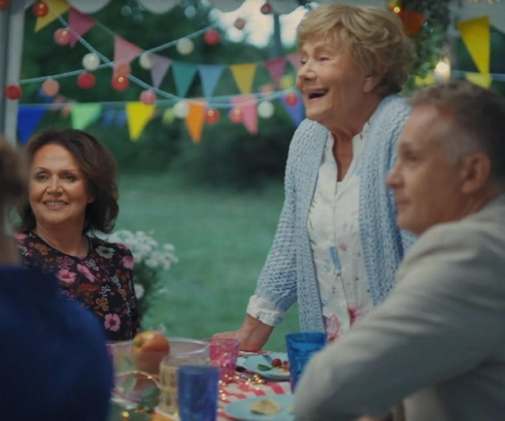 Uśmiechnięta Barbara Mostowiak (Teresa Lipowska) w niebieskim swetrze, oparta o stół z jedzeniem, rozmawia z gośćmi na przyjęciu w ogrodzie. Na stole kolorowe naczynia, jedzenie i dekoracje. W tle zielony ogród i dekoracje z okazji jubileuszu M jak miłość. Więcej o tym wydarzeniu znajdziesz na portalu Super Seriale.