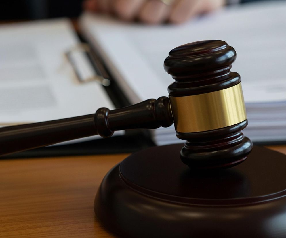 Na pierwszym planie, z prawej strony, znajduje się drewniany młotek sędziowski w kolorze ciemnobrązowym, z błyszczącą, złotą obręczą wokół głowicy. Młotek opiera się na dopasowanej, okrągłej podstawce w tym samym kolorze. W tle, po lewej stronie, widoczne są dłonie osoby trzymającej długopis, pochylonej nad otwartymi dokumentami, które leżą na teczce lub podkładce. Obraz utrzymany jest w ciepłej tonacji, z brązowym drewnianym blatem stołu i białymi kartkami papieru.