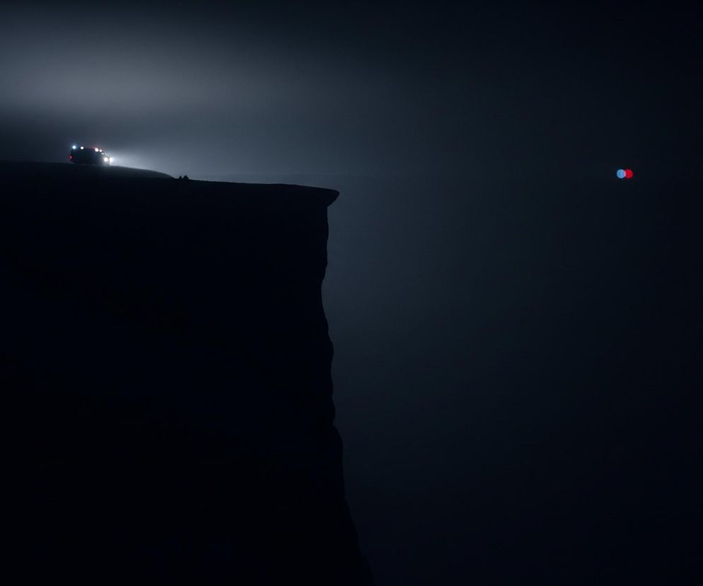 Ciemna sylwetka klifu dominuje lewą część obrazu, ostro kończąc się u góry. Na jego krawędzi widać zarys pojazdu terenowego o włączonych reflektorach, które emitują silne, białe światło, rozpraszające się w zamglonym otoczeniu. Nieco za pojazdem dostrzegalna jest sylwetka człowieka. Na ciemnym, rozmytym tle, po prawej stronie zdjęcia, unoszą się dwie rozmyte kule światła – jedna czerwona i jedna niebieska, ułożone poziomo obok siebie.