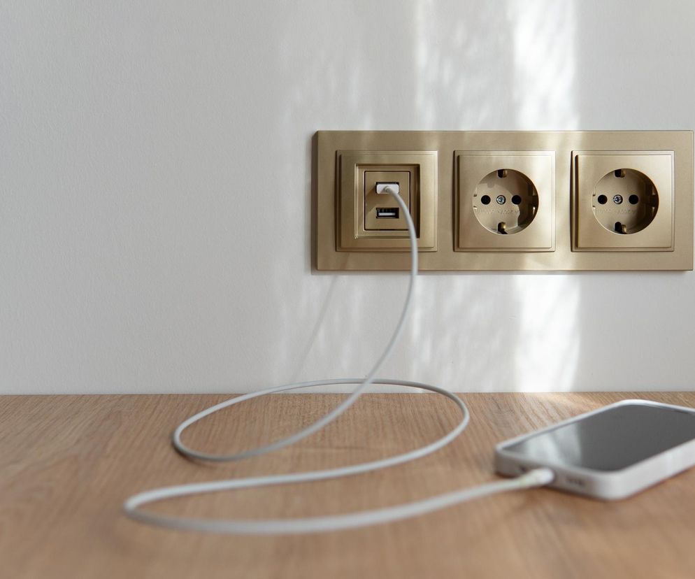 Smartfon podłączony do gniazdka z ładowarką USB i dwoma gniazdami sieciowymi na ścianie. Ilustracja symbolizuje zagrożenie poborem prądu przez urządzenia w trybie czuwania i ryzyko awarii, o czym przeczytasz na Super Biznes.