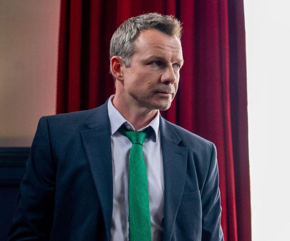 Aktor Lesław Żurek jako Bruno w granatowej marynarce, białej koszuli i zielonym krawacie, z włosami zaczesanymi do góry, na tle czerwonej zasłony i jasnej ściany. Aktor zastanawia się i patrzy w bok. Więcej o losach jego postaci w serialu Barwy szczęścia przeczytasz na stronie Super Seriale.