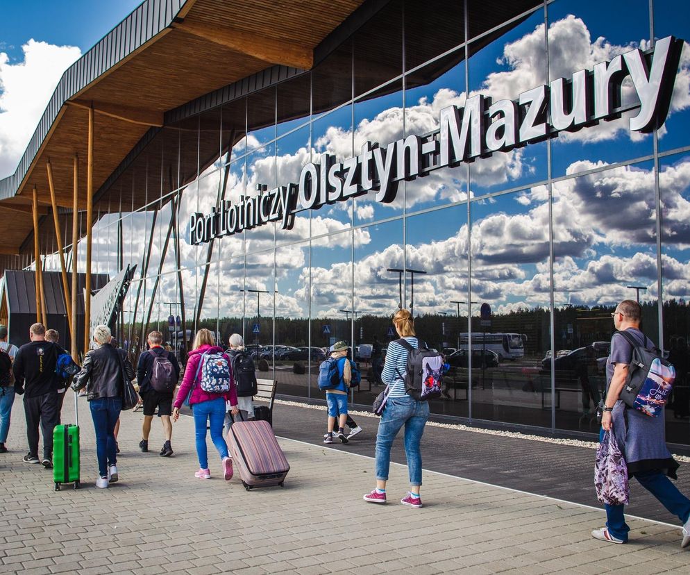 Pasażerowie z bagażami przed nowoczesnym terminalem lotniska Olsztyn-Mazury, którego szklana fasada odbija błękitne niebo z chmurami, symbolizując nadchodzące zmiany w podróżowaniu. Więcej o wymogach ETA przeczytasz na Super Biznes.