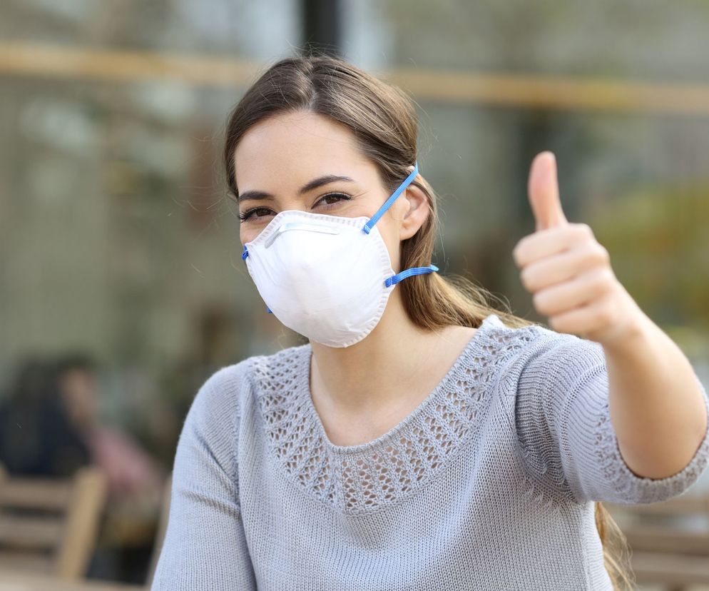 Kobieta w białej maseczce ochronnej typu N95, z brązowymi włosami rozpuszczonymi na plecach, ubrana w jasnoszary sweter, pokazuje kciuk w górę, symbolizując bezpieczeństwo i skuteczność maseczek domowej roboty. Więcej na ten temat przeczytasz na Poradnik Zdrowie.