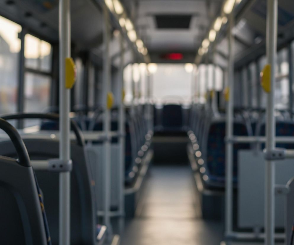 Wnętrze autobusu widoczne jest z perspektywy siedzącego pasażera, z rozmazanym tłem. Przednie oparcia siedzeń znajdują się na pierwszym planie po lewej i prawej stronie, w kolorze ciemnoszarym, z uchwytami w tym samym kolorze. W oddali, wzdłuż obu stron pojazdu, ciągnie się rząd identycznych siedzeń z niebieskimi, wzorzystymi obiciami. Od sufitu do podłogi biegną pionowe, jasnoszare słupki z żółtymi przyciskami do zatrzymywania autobusu, widocznymi po lewej stronie. Światło dzienne wpada przez liczne okna, które ukazują niewyraźny krajobraz miejski. Podłoga jest jasna, a sufit ciemniejszy, z rzędem małych, jasnych świateł biegnących przez całą długość pojazdu.