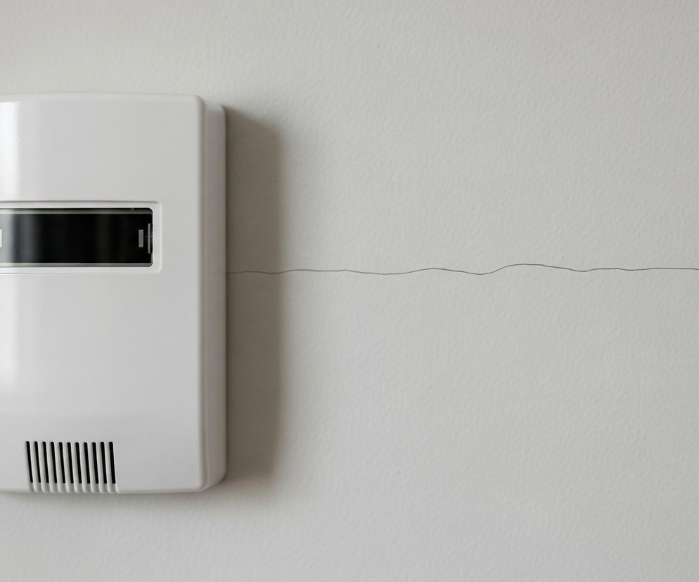 Na jasnoszarej, teksturowanej ścianie po lewej stronie zamontowane jest białe prostokątne urządzenie. Na jego froncie znajduje się poziomy, czarny wyświetlacz z jasnym, pionowym paskiem po lewej stronie oraz kratka wentylacyjna u dołu. Od prawej krawędzi urządzenia w kierunku prawej strony zdjęcia biegnie cienka, ciemna, falista linia.