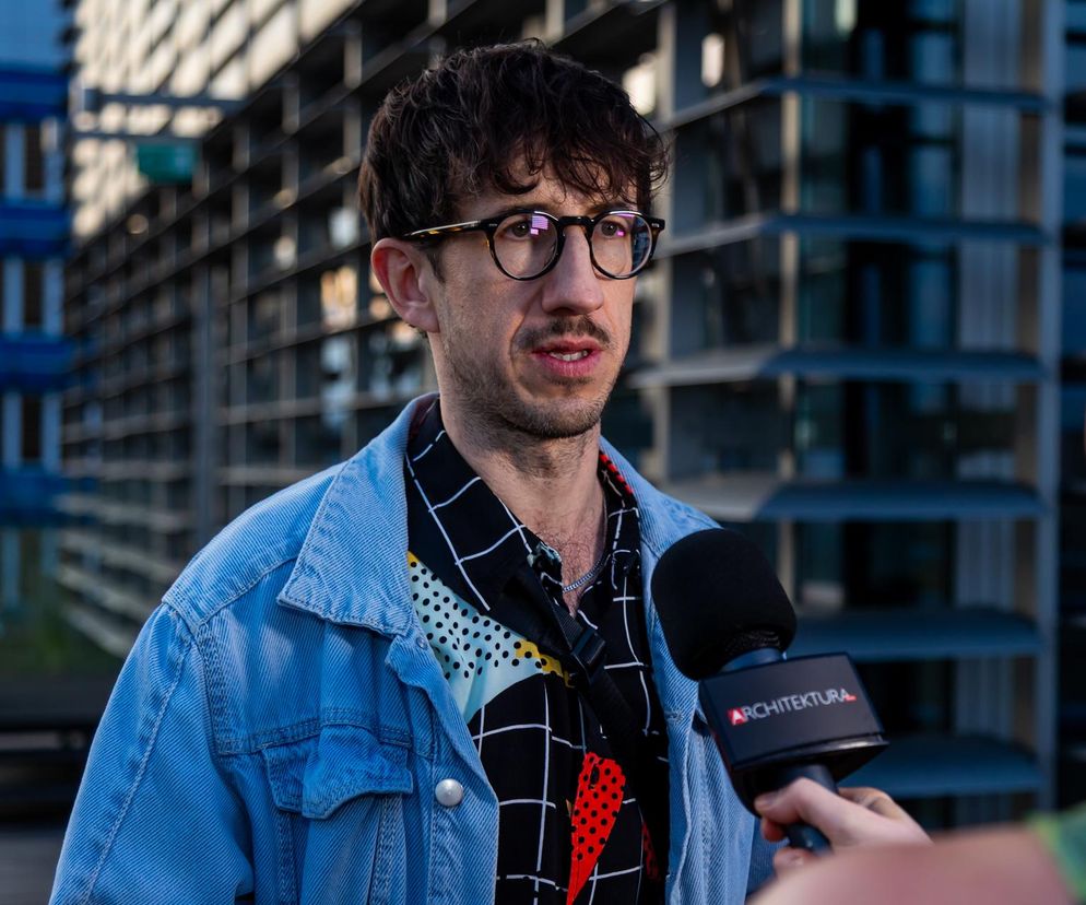 Grzegorz Piątek, architekt, krytyk i historyk architektury, udziela wywiadu, trzymając mikrofon z logo Architektura. Na sobie ma jasnoniebieską jeansową kurtkę i wzorzystą koszulę, w tle widać nowoczesny budynek. O osiągnięciach i twórczości Grzegorza Piątka można przeczytać na Architektura Murator Plus.