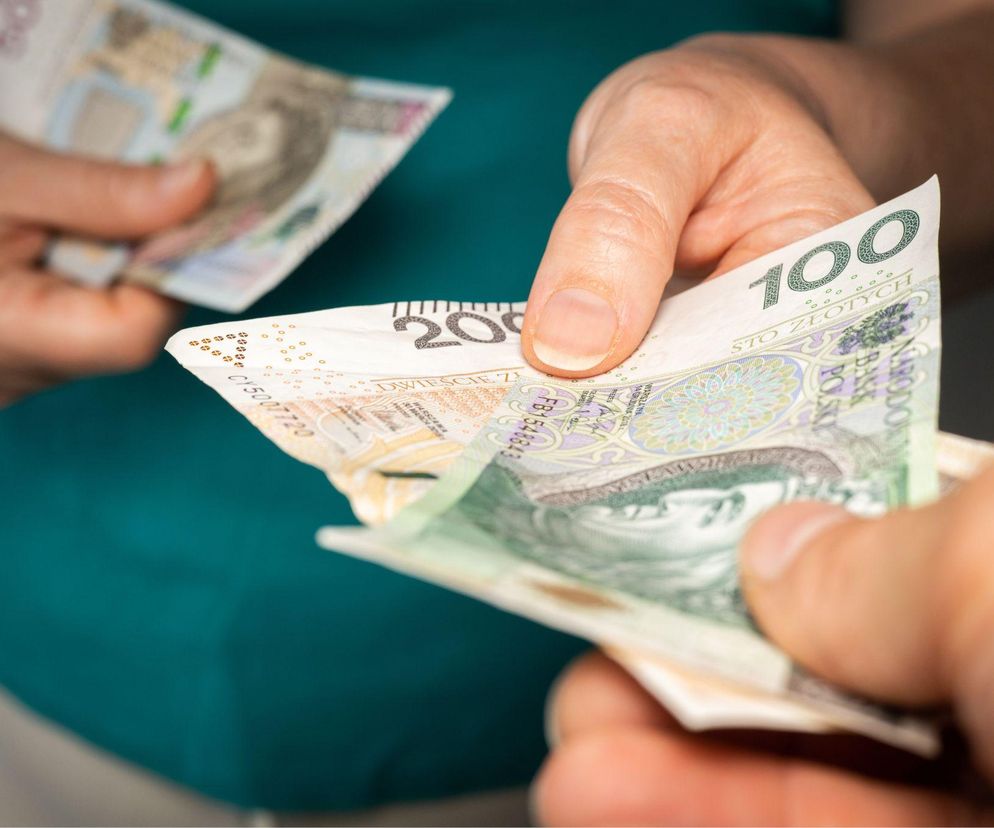 Dłonie dwóch osób, z których jedna przekazuje banknoty studwudziestozłotowe drugiej, symbolizując świadczenia wspierające dla osób z niepełnosprawnościami. Dowiedz się więcej o warunkach ich otrzymania na Poradnik Zdrowie.