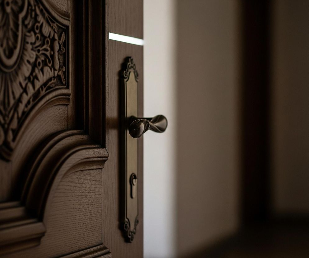 Otworzone drzwi ukazują ciemne wnętrze pokoju. Lewa część drzwi jest brązowa, drewniana, z bogatymi rzeźbieniami roślinnymi i wzorami na powierzchni. Klamka jest metalowa, w kolorze starego złota, z dekoracyjnym, podłużnym szyldem. Po prawej stronie, za otwartymi drzwiami, widać rozmazany, jasny fragment ściany i ciemniejszą przestrzeń pokoju.