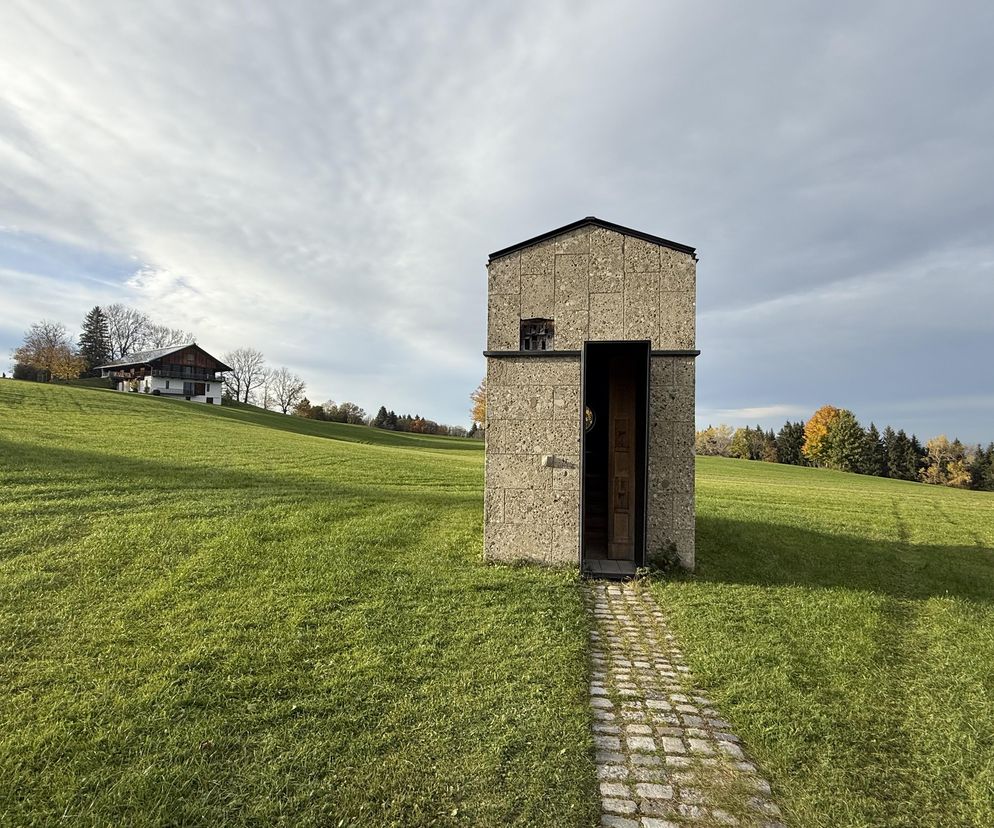 Kapliczka Michele de Lucchi w Alpach Bawarskich, wykonana z lokalnego kamienia wapiennego z drewnianymi drzwiami, prowadzi do niej ścieżka z kostki brukowej. W tle zielone wzgórza z nielicznymi budynkami i drzewami, pod zachmurzonym niebem. Więcej o minimalistycznej architekturze sakralnej przeczytasz na Architektura Murator Plus.