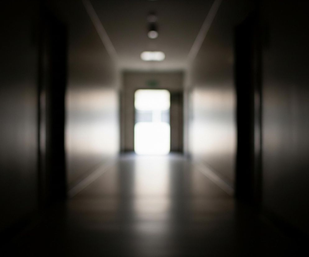 Długi korytarz, ciemny po bokach, rozświetla się w oddali, gdzie widać jasne, białe światło wydobywające się zza otwartych drzwi. Oświetlona jest również podłoga i część ścian w głębi, tworząc refleksy i rozmyte kształty. Całość obrazu jest nieostra, z dominującymi odcieniami szarości i czerni, a jasne elementy są mocno prześwietlone.