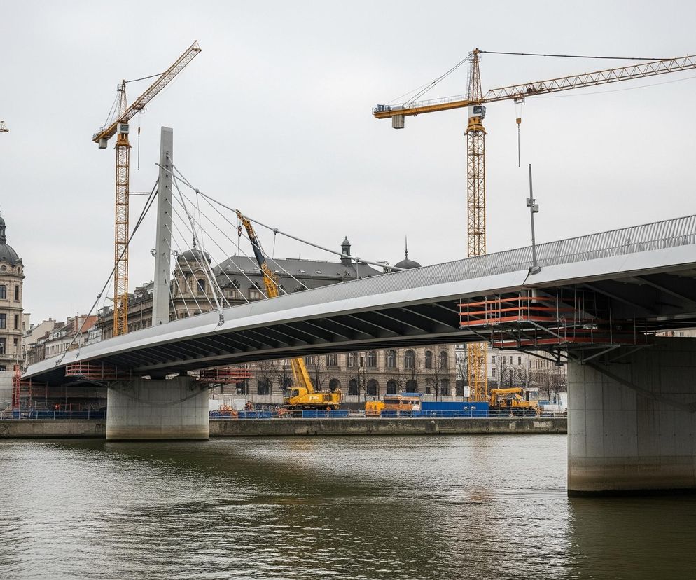 Duży, nowoczesny most o jasnoszarej konstrukcji i białych barierkach biegnie przez środek kadru, wsparty na solidnych betonowych filarach zanurzonych w brudnozielonej wodzie rzeki. Nad i wokół mostu widoczne są liczne żółte dźwigi budowlane o kratownicowej konstrukcji, co wskazuje na trwające prace. Na lewym brzegu rzeki wznosi się imponujący, beżowy budynek z kopułą, przypominający klasyczną architekturę, a za nim kolejny, mniejszy budynek z kopułą; oba sąsiadują z placem budowy, na którym stoją niebieskie kontenery i kolejne żółte maszyny budowlane. Niebo jest jednolicie szare, co nadaje scenie stonowany charakter.