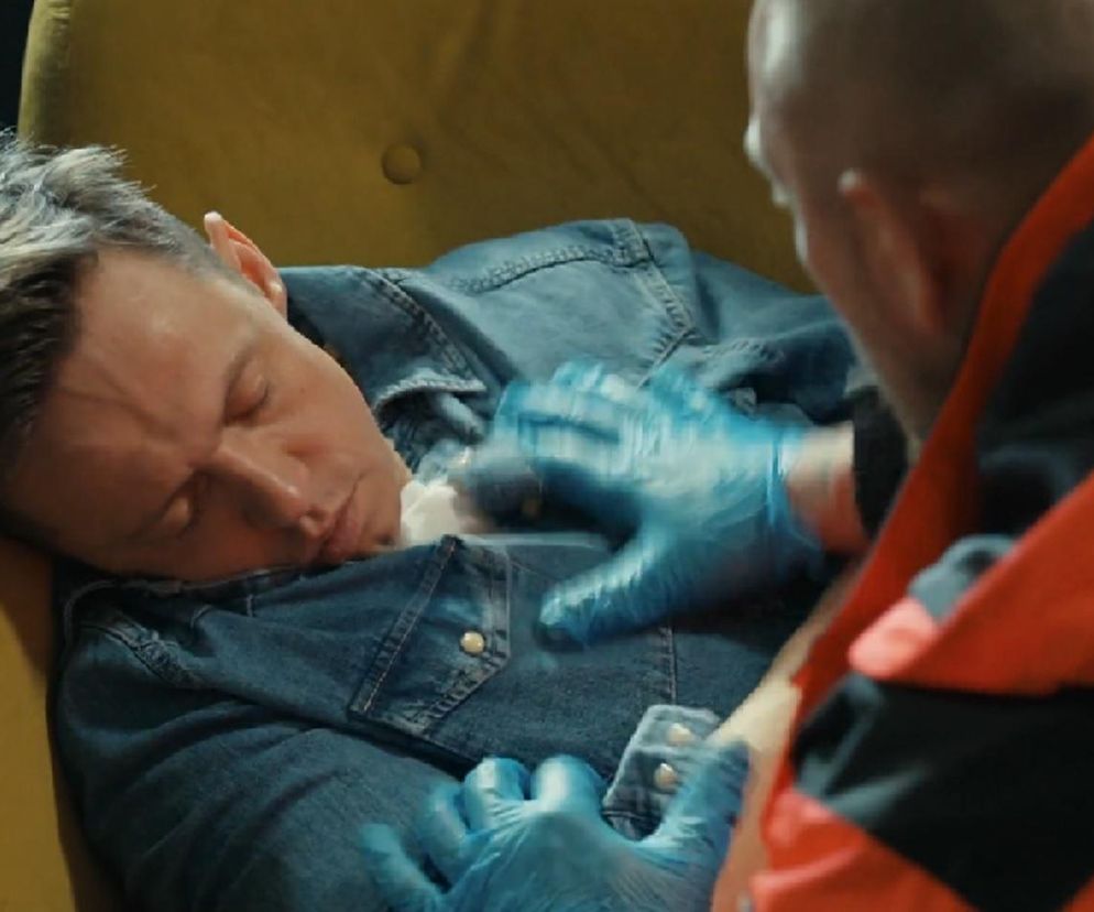 Piotr Ligienza jako Błażej Modrzycki leży nieprzytomny na żółtym fotelu, z białą chusteczką na koszuli. Ratownik medyczny w niebieskich rękawiczkach bada mu puls, a to wszystko możesz przeczytać w artykule Super Seriale.