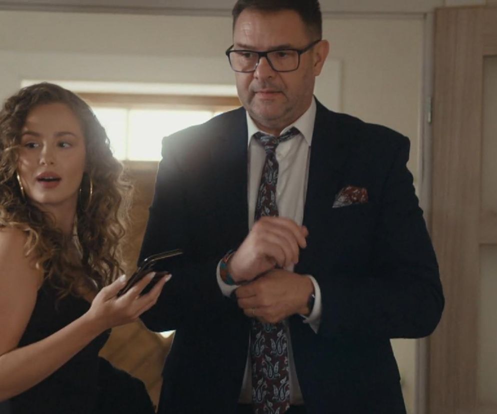 Tomasz Karolak jako Ludwik Boski w garniturze i Wiktoria Gąsiewska jako Agata w czarnej sukience, trzymająca telefon, rozmawiający w scenie z serialu Rodzinka.pl. Więcej informacji o serialach znajdziesz na Super Seriale.