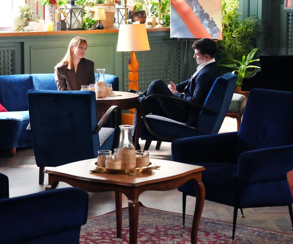 Michalina Robakiewicz (Ola) i Jasper Sołtysiewicz (Justin) siedzą naprzeciwko siebie w kawiarni, rozmawiając przy stole. Ona w beżowej marynarce na granatowej sofie, on w ciemnym garniturze w fotelu, co sugeruje kluczowy moment z serialu Barwy szczęścia, o którym przeczytasz więcej na Super Seriale.