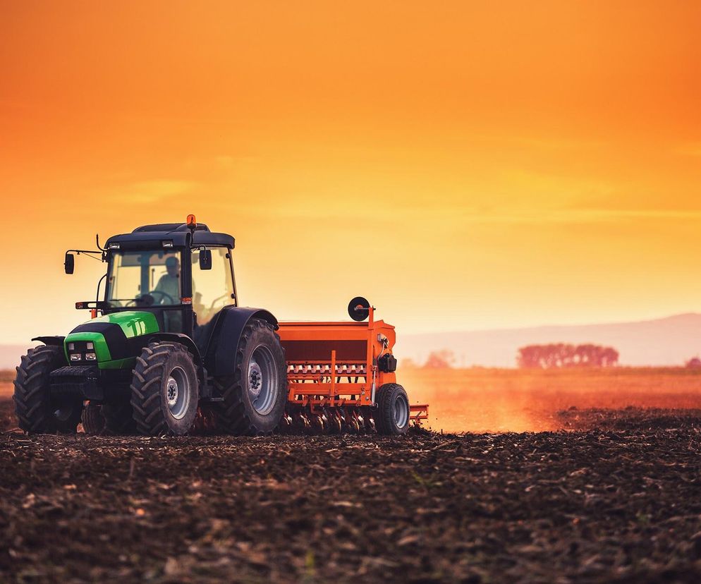 Zielony traktor ciągnący pomarańczowy siewnik na zaoranym polu, przygotowującym ziemię pod uprawy. Maszyny rolnicze o zmierzchu. Analizę ryzyka wypadków w rolnictwie, szczególnie w mniejszych gospodarstwach, znajdziesz na Super Biznes.