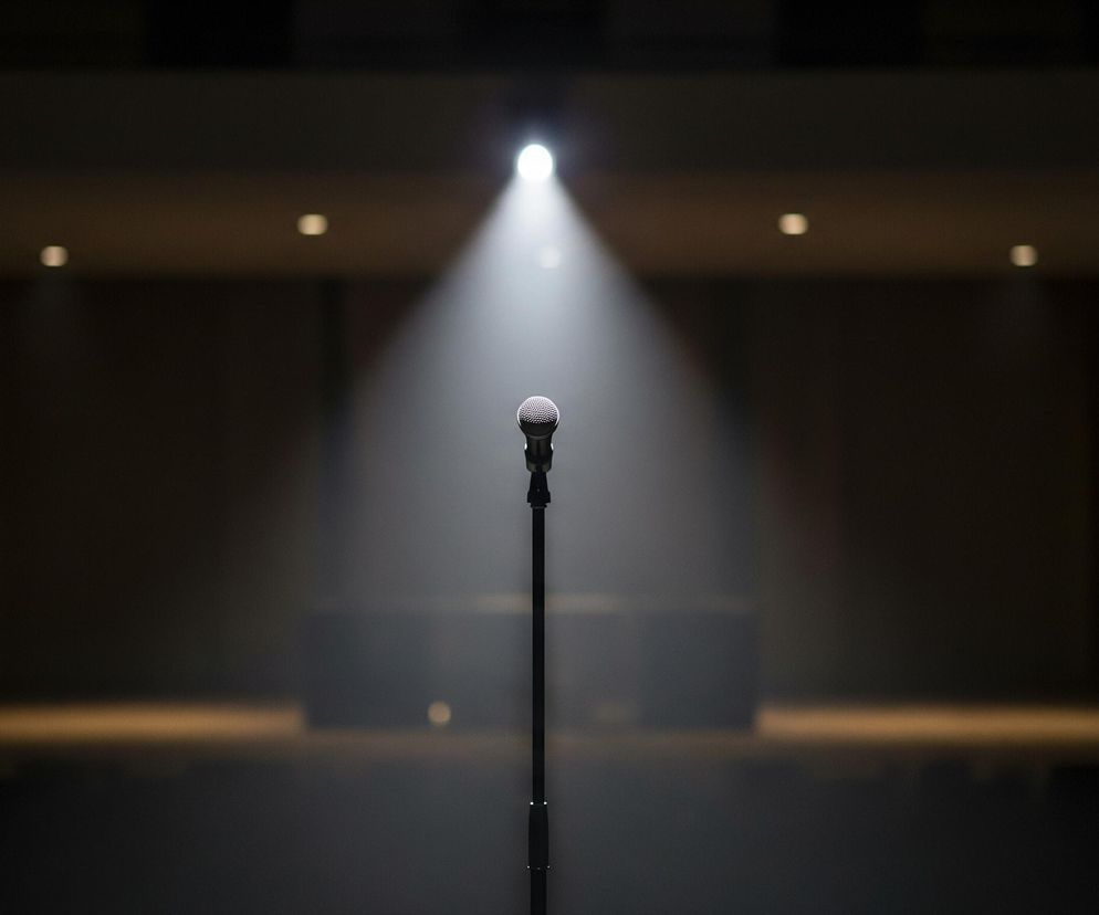 Na pierwszym planie, centralnie, stoi czarny mikrofon na stojaku, wyraźnie oświetlony mocnym, białym snopem światła padającym z góry. W tle widać ciemną przestrzeń, przypominającą widownię lub salę koncertową, z lekko rozmazanymi, niewyraźnymi elementami brązowo-szarych ścian i kilkoma słabymi punktami światła widocznymi na suficie. Scena jest skąpana w mroku, z wyjątkiem intensywnie podświetlonego miejsca mikrofonu, co tworzy silny kontrast.