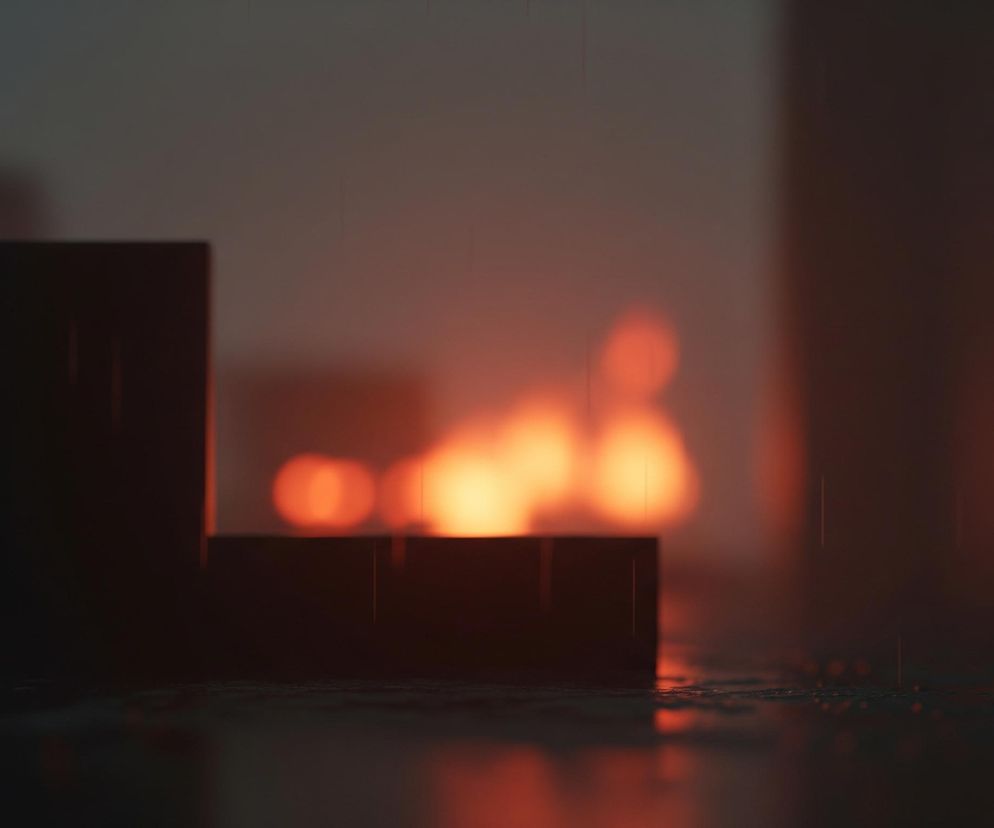 Widoczna jest ciemna, deszczowa scena z rozmyśloną głębią ostrości. Na pierwszym planie po lewej stronie znajdują się dwa czarne, prostokątne obiekty o schodkowym kształcie, na których powierzchni widać odbicia światła i pojedyncze, cienkie linie symbolizujące krople deszczu. W tle dominują rozmyte, pomarańczowe i żółte plamy światła, prawdopodobnie pochodzące od źródeł ognia lub intensywnych lamp, które rzucają ciepłą poświatę na całą kompozycję. Całość jest ciemna i nastrojowa, z wyraźnym efektem deszczu padającego na wilgotne powierzchnie.