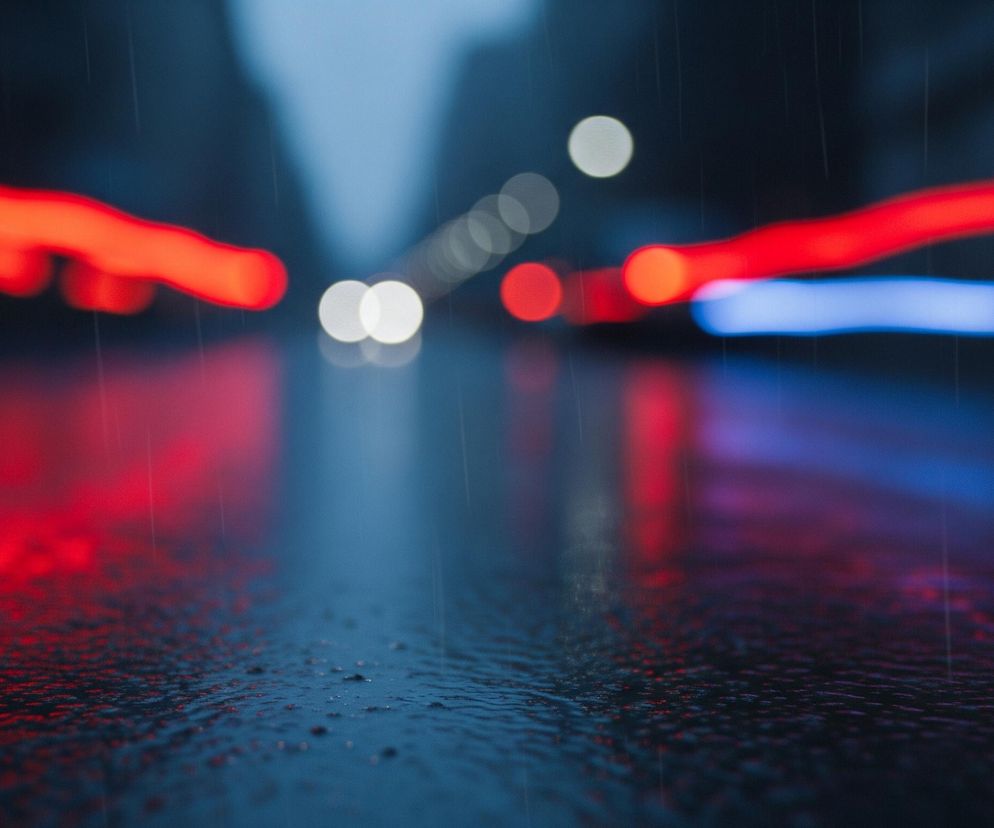Ciemną, deszczową ulicę z rozmytymi światłami w oddali. Na pierwszym planie widoczna jest mokra powierzchnia asfaltu, odbijająca czerwone i niebieskie smugi światła, które zdają się płynąć po jezdni. W tle, rozmyte jasne i czerwone kule światła tworzą wrażenie głębi i ruchu, a subtelne, pionowe linie sugerują padający deszcz. Dominują ciemne, chłodne odcienie niebieskiego i szarości, kontrastujące z jaskrawą czerwienią świateł.