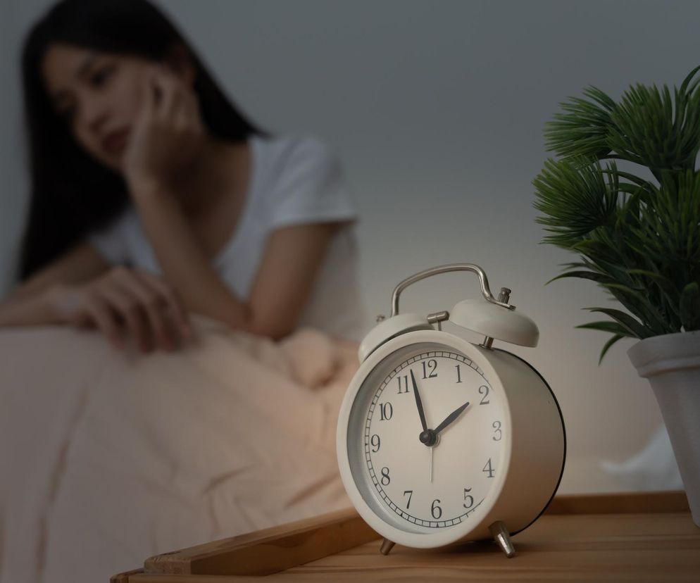 Biały budzik na stoliku nocnym, wskazujący godzinę drugą, przed którym siedzi smutna, młoda kobieta z bezsennymi oczami, z niewyraźną miną w tle, w półmroku, symbolizujący problemy ze snem i ryzyko demencji, o których możesz przeczytać na portalu Poradnik Zdrowie.