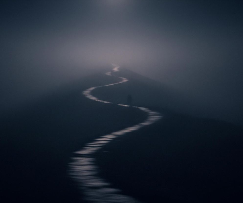 Długa, kręta ścieżka przecina ciemny, zamglony krajobraz, wijąc się od dolnej części zdjęcia ku górze. Światło, jaśniejsze w górnej części obrazu, tworzy blask rozświetlający ścieżkę i jej najbliższe otoczenie, stopniowo zanikając w ciemność po bokach i na dole. Na ścieżce, mniej więcej w połowie długości, stoi mała, niewyraźna sylwetka człowieka, otoczona mgłą.