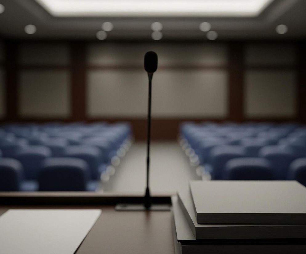 Na pierwszym planie znajduje się ciemnobrązowe, drewniane podium z czarnym mikrofonem na długiej nóżce, stojącym centralnie. Po lewej stronie mikrofonu leży pojedyncza biała kartka papieru, a po prawej widoczny jest stos czterech zamkniętych, szarych teczek lub książek. Tło rozmywa się w głąb pomieszczenia, gdzie widać rzędy pustych, niebieskich foteli ustawionych w sali, która ma jasne, kasetonowe ściany z ciemnymi drewnianymi ramami. Na suficie rozmieszczone są jasne, punktowe źródła światła, a centralnie znajduje się prostokątne, podłużne oświetlenie.