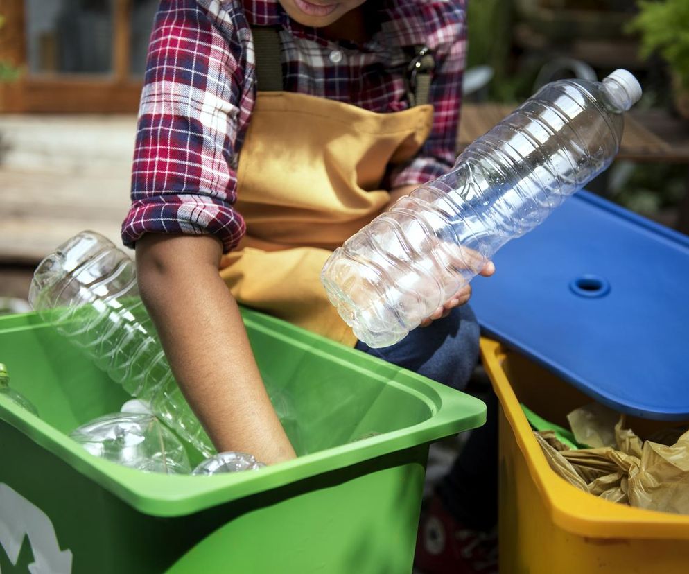 Osoba w kraciastej koszuli i żółtym fartuchu segreguje plastikowe butelki do zielonego pojemnika na tle żółtego pojemnika z papierem. Gminy rezygnują z darmowych worków na śmieci, wprowadzając nowe zasady. Przeczytaj więcej o zmianach w segregacji odpadów i karach za nieprawidłowe sortowanie na Super Biznes.