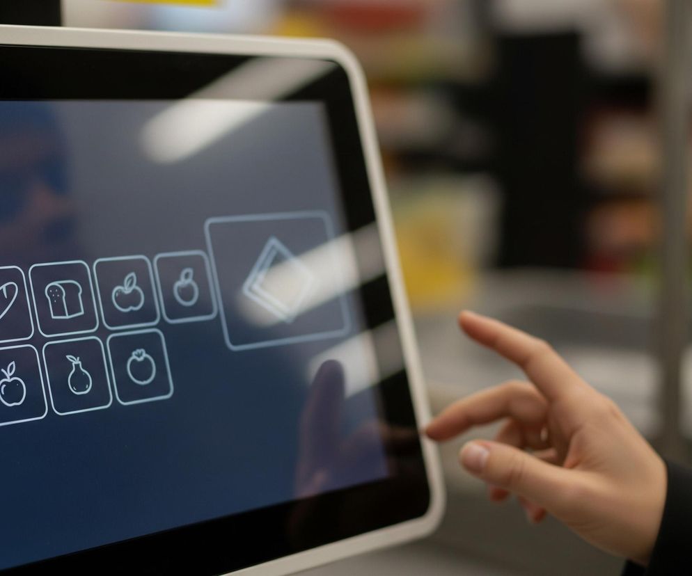 Blisko kadru, po prawej stronie, jasna dłoń dotyka ekranu dotykowego z menu produktów. Na ciemnym ekranie widoczne są białe, liniowe ikony, przedstawiające m.in. bochenki chleba, kromki chleba tostowego, jabłka, gruszki i pomidora, rozmieszczone w dwóch rzędach. W tle, po prawej, znajduje się rozmyty element kasy samoobsługowej w jasnym kolorze, a dalej widać niewyraźne półki sklepowe.