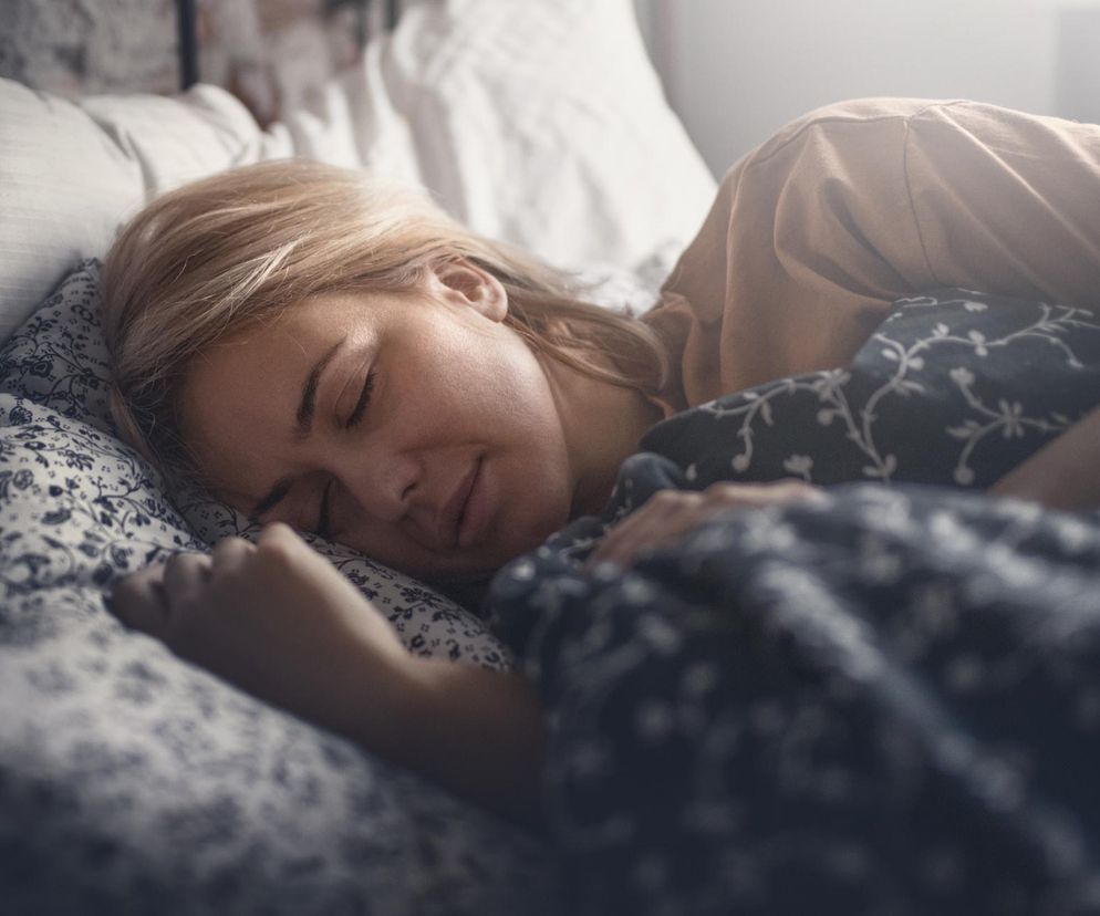 Kobieta o blond włosach, ubrana w beżową koszulkę, śpi spokojnie w łóżku, otulona wzorzystą kołdrą. Jej twarz jest zrelaksowana, oczy zamknięte, a lekki uśmiech świadczy o głębokim śnie, który, jak czytamy na Poradnik Zdrowie, ma kluczowe znaczenie dla pamięci.