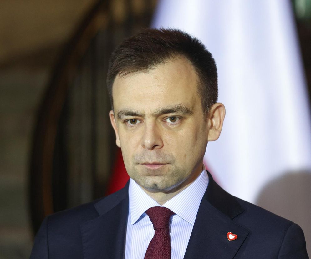 Portret Andrzeja Domańskiego, ministra finansów, w granatowym garniturze z przypinką serca, jasną koszuli w paski i bordowym krawacie. Tło częściowo rozmyte, widoczna biała flaga. Artykuł o oszczędnościach budżetu państwa, o czym więcej przeczytasz na Super Biznes.