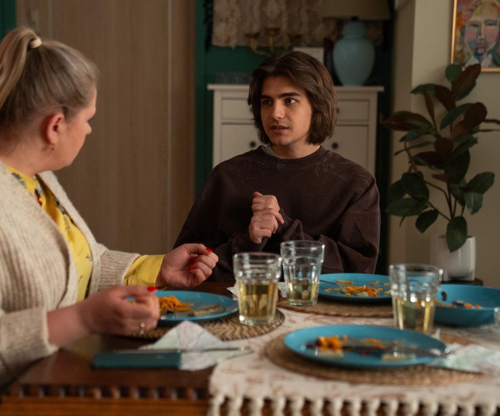 Młody Romeo (Jacek Tyszkiewicz) siedzi przy stole, rozmawiając z Aldoną (Elżbieta Romanowska) z serialu Barwy szczęścia. Na stole niebieskie talerze, szklanki z napojem. Romeo wygląda na zamyślonego, co możesz przeczytać na Super Seriale.